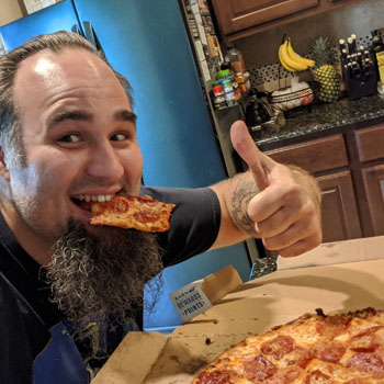 Josh Wolfer flashes a thumbs up with a slice of pizza in his mouth.