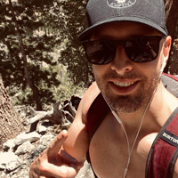 Scott Davidson, shirtless, smiles to the camera while hiking.